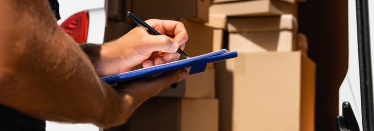Pessoa fazendo anotações em uma prancheta em frente a uma van carregada com caixas, representando o controle e cálculo do giro de estoque em operações logísticas e de distribuição.