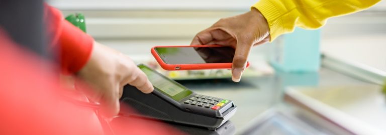 Cliente segurando um celular para realizar um pagamento por aproximação em uma máquina de cartão (POS) operada por um funcionário. A imagem ilustra uma transação moderna no Ponto de Venda (PDV) de uma loja.