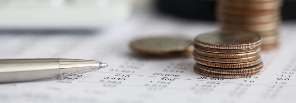 Uma foto em primeiro plano de uma caneta prateada apontando para uma planilha de números. No lado direito, há uma pilha de moedas empilhadas sobre a planilha.