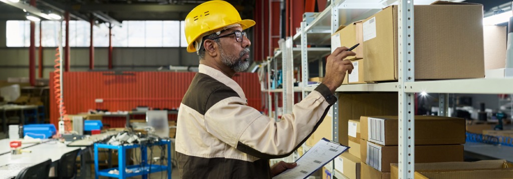Funcionário em armazém organizando o estoque físico, conferindo caixas nas prateleiras com prancheta e caneta, em ambiente logístico.
