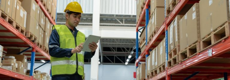Funcionário em centro de distribuição usando tablet para conferência de mercadorias, entre prateleiras com caixas, representando ações de prevenção de perdas no estoque.