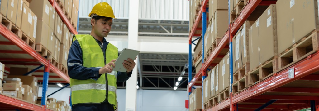 Funcionário em centro de distribuição usando tablet para conferência de mercadorias, entre prateleiras com caixas, representando ações de prevenção de perdas no estoque.